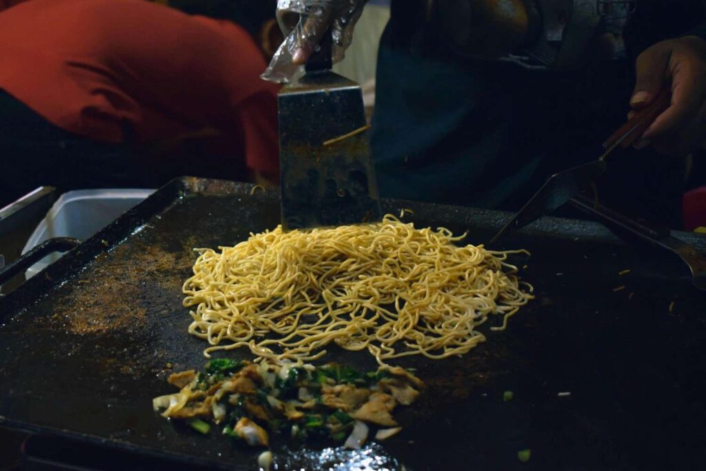 A cook uses metal spatulas to manage a large pile of yellow noodles on a dark, flat-top griddle. Separated from the pasta, a small portion of chopped meat and green vegetables sizzles on the hot surface, suggesting a street food preparation setting.