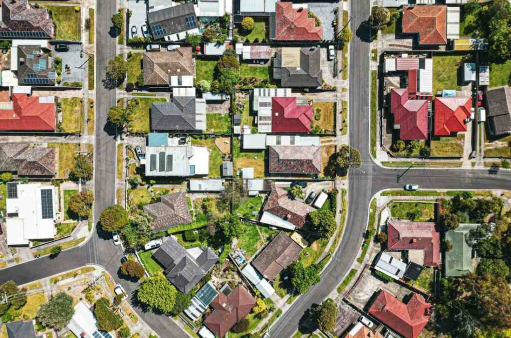 This high-angle aerial photograph captures a vibrant suburban neighborhood characterized by a geometric patchwork of homes with varying red, grey, and white rooftops. Winding asphalt streets cut through the residential blocks, revealing details like backyard gardens, parked cars, and solar panels atop several houses.