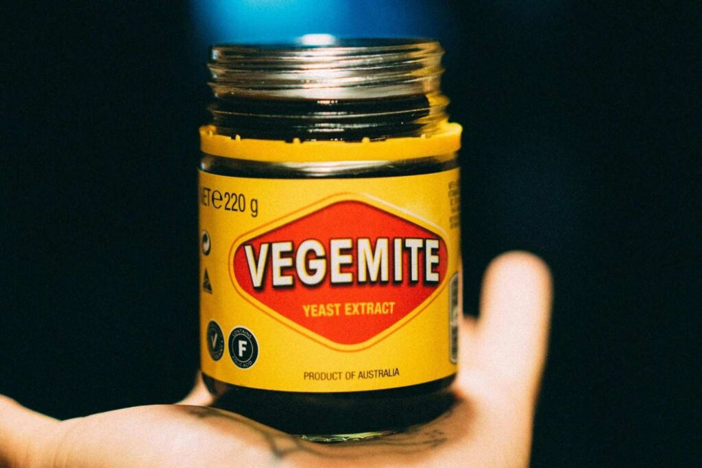 A person's open palm cradles a jar of Vegemite, clearly displaying the iconic bright yellow label with the red diamond logo and bold white text. The close-up shot utilizes a shallow depth of field, keeping the Australian yeast extract spread in sharp focus while blurring the hand and the dark background.