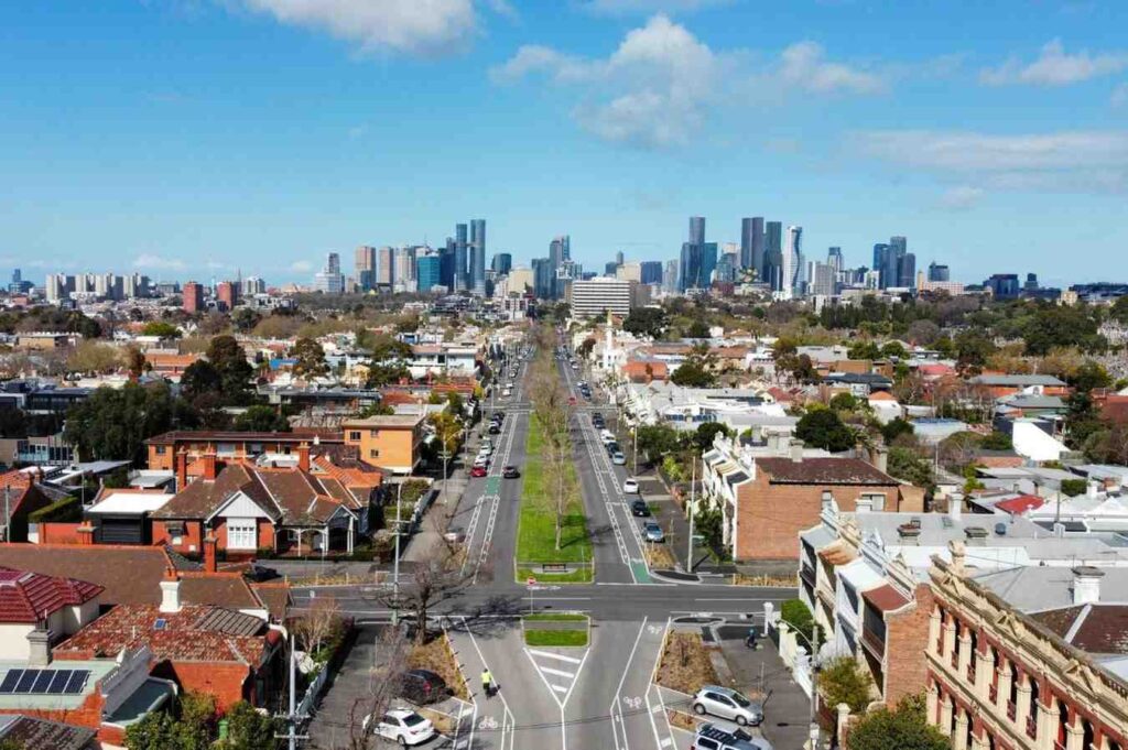 This wide aerial shot captures a straight, tree-lined boulevard running through a district of red-roofed homes directly towards a dense, modern city skyline. The composition highlights the architectural transition from the sprawling, low-rise suburbs in the foreground to the towering skyscrapers in the distance under a partly cloudy sky.