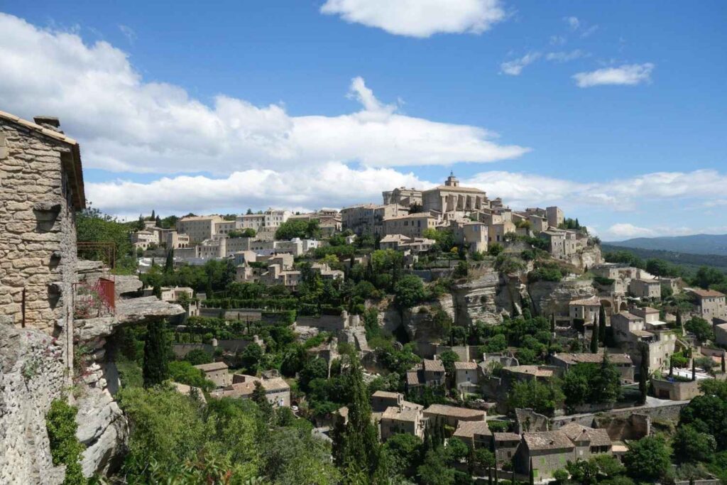 A historic stone village with terracotta-roofed buildings is perched dramatically on a rugged, terraced hillside. The town is surrounded by dense green vegetation and steep rocky cliffs, all set under a bright blue sky filled with fluffy white clouds.