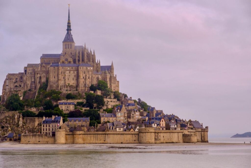 The majestic Mont-Saint-Michel abbey rises from a rocky island, topped by a slender spire that pierces the hazy, lavender-colored sky. A dense cluster of historic stone buildings and defensive walls sits at its base, meeting the wet sands of the surrounding tidal bay at low tide.