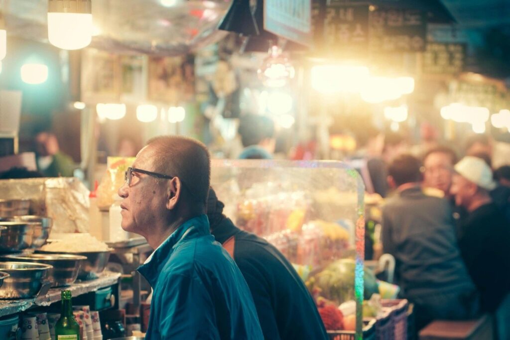 This photo captures an older man wearing glasses and a blue jacket sitting contemplatively at a street food counter. The background is filled with the warm glow of market lights and a blurred crowd of diners, creating a lively nighttime atmosphere.