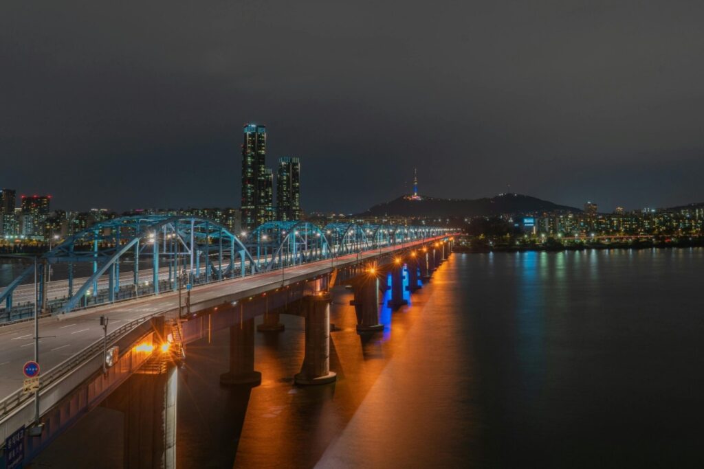 This nightscape captures a long bridge with illuminated blue arches spanning a wide river, where warm orange streetlights reflect vividly against the dark water. In the background, modern high-rise buildings stand on the left, while the iconic N Seoul Tower glows atop a distant hill against the dark sky.