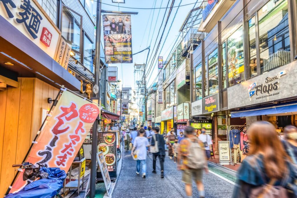 This vibrant street in Shimokitazawa is filled with a steady flow of pedestrians moving past a mix of modern storefronts, including a used clothing shop and various restaurants. Bright banners and vertical signs line the narrow walkway, while a complex network of power lines crisscrosses the sky above the bustling neighborhood.