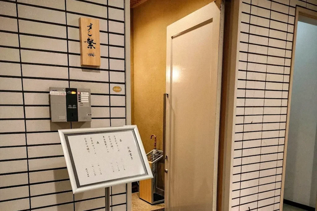 Entrance to a Japanese-style sushi restaurant with a white grid-patterned wall, wooden sign with Japanese characters, menu board, and a partially open door.