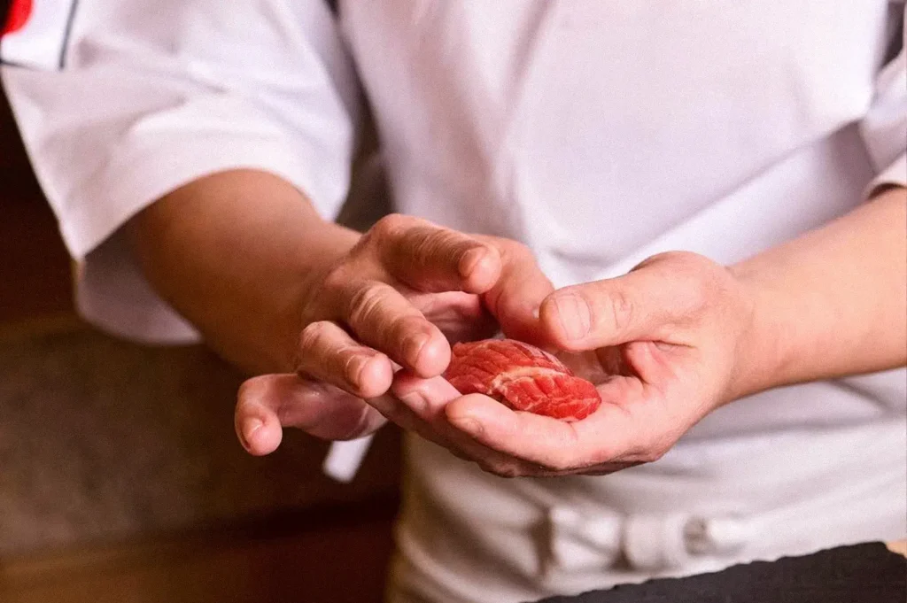 Close-up of a sushi chef shaping a small piece of marbled red fish with their hands.