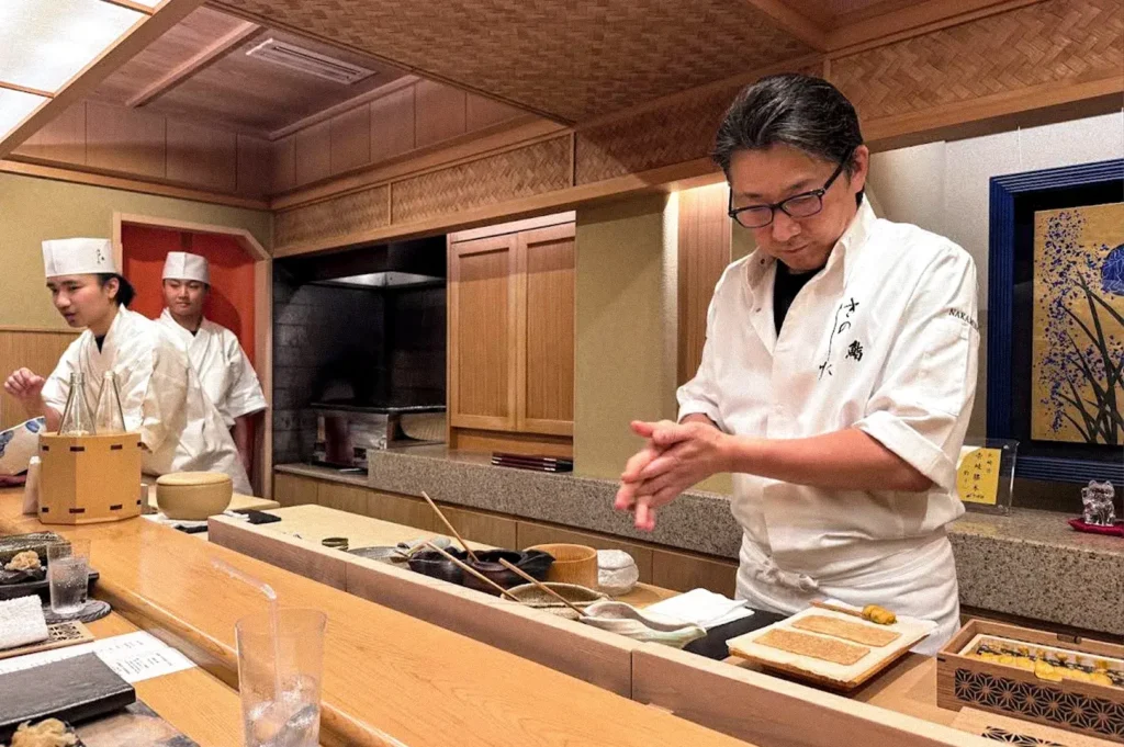 Sushi chef in a white uniform preparing food behind a wooden counter, with two other chefs working in the background.
