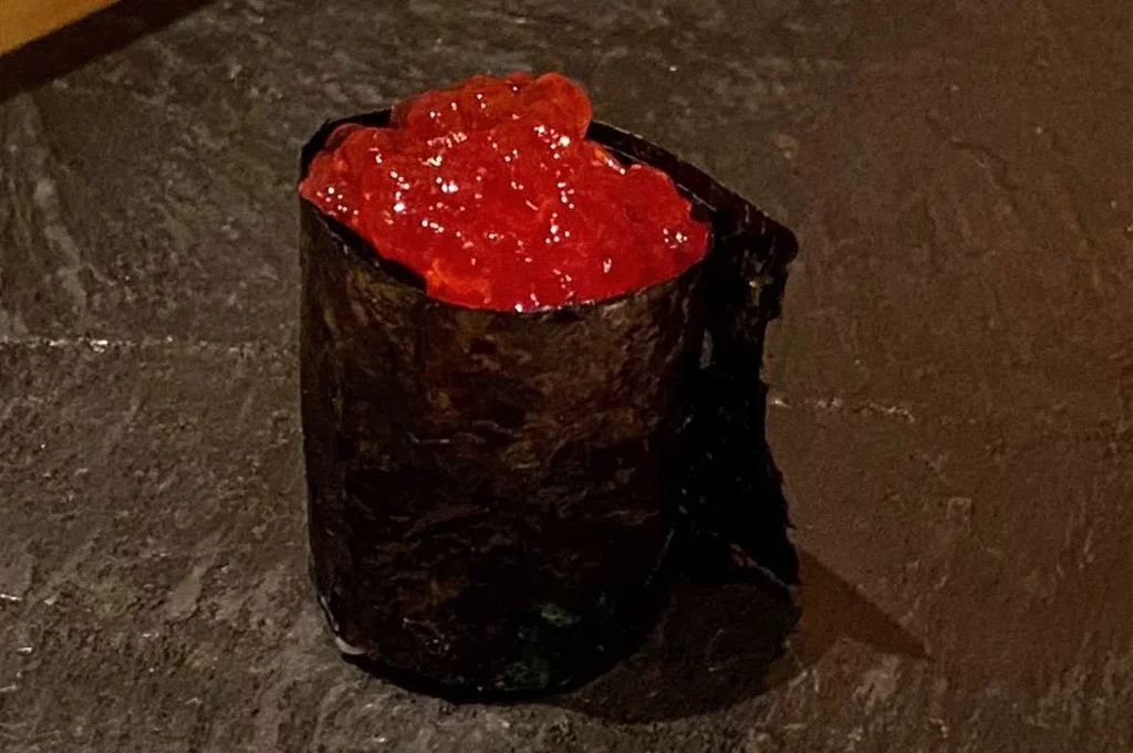 Close-up of a glossy red-topped sushi piece wrapped in dark seaweed and served on a dark counter.
