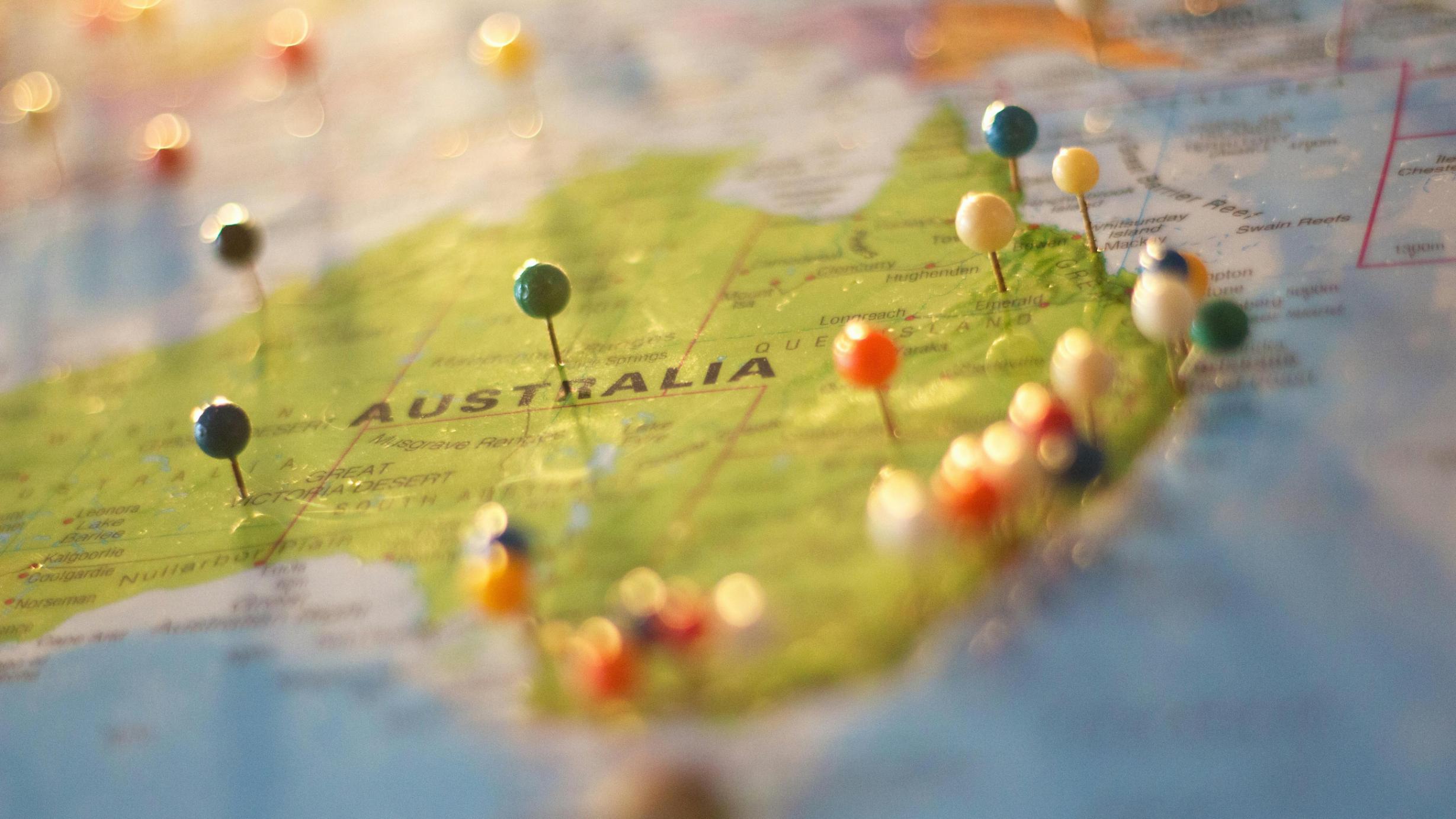 This is a close-up, shallow depth-of-field shot of a world map centered on the continent of Australia, which is marked with several brightly colored pushpins.