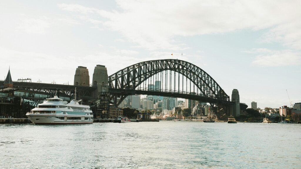 This image showcases the iconic Sydney Harbour Bridge with a large, white cruise ship or ferry docked prominently on the left side of the frame.