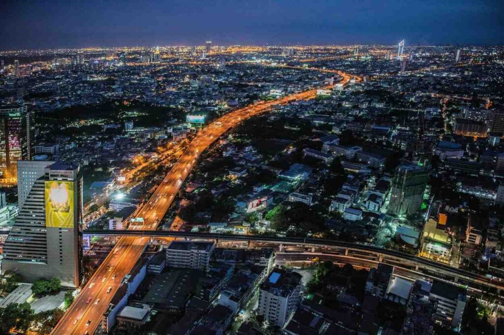 An aerial night view captures a sprawling metropolis, dominated by a winding expressway that cuts through the dark cityscape like a river of glowing orange light. Surrounding the highway, dense clusters of buildings and skyscrapers stretch toward the horizon, filling the landscape with thousands of shimmering city lights.