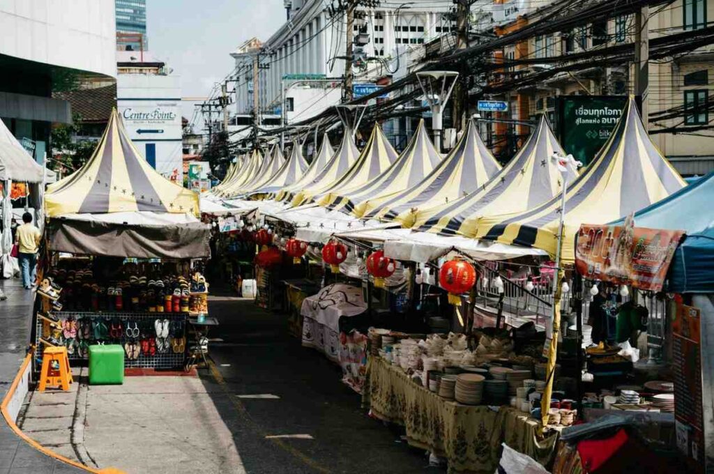 A bustling street market features a uniform row of tents with yellow and gray striped peaked roofs, sheltering stalls filled with ceramic dishware and racks of footwear. The vibrant scene is set against a backdrop of city buildings and tangled overhead power lines, with red lanterns hanging beneath the canopies to add a touch of color to the lively walkway.