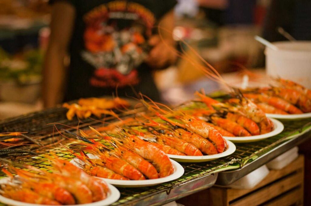 Rows of white plates are lined up on a metal rack, each filled with large, bright orange grilled prawns that feature long antennae and charred shells.
