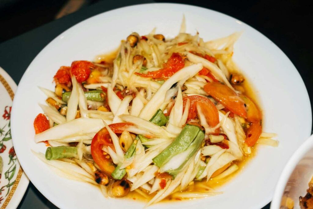 A white plate presents a vibrant serving of Som Tum (green papaya salad), featuring shredded papaya tossed with red tomato slices, green beans, and roasted peanuts. The dish is coated in a glossy dressing that highlights the fresh, crisp textures of the ingredients, creating an appetizing close-up of this classic Thai street food.