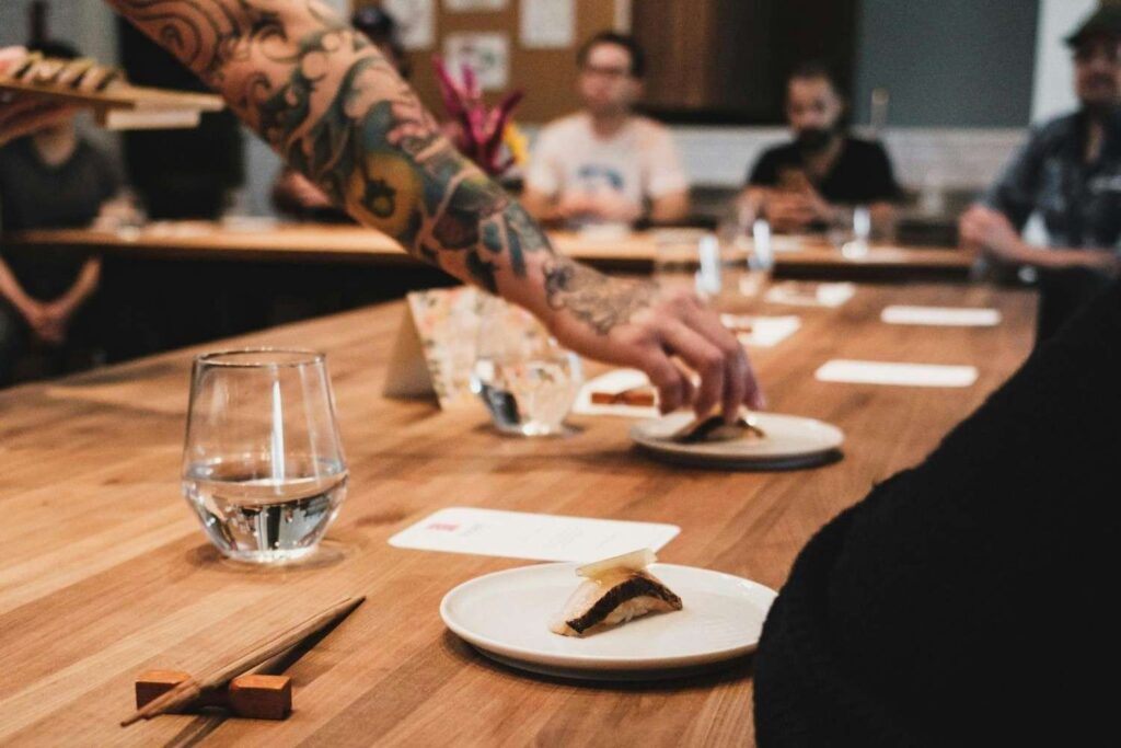 This close-up shot captures a communal dining scene where a person with a heavily tattooed arm reaches across a wooden table to place a small plate of food. In the sharp foreground, a piece of sushi rests near chopsticks and a glass of water, while blurred diners sit in the background, evoking an intimate and casual culinary atmosphere.