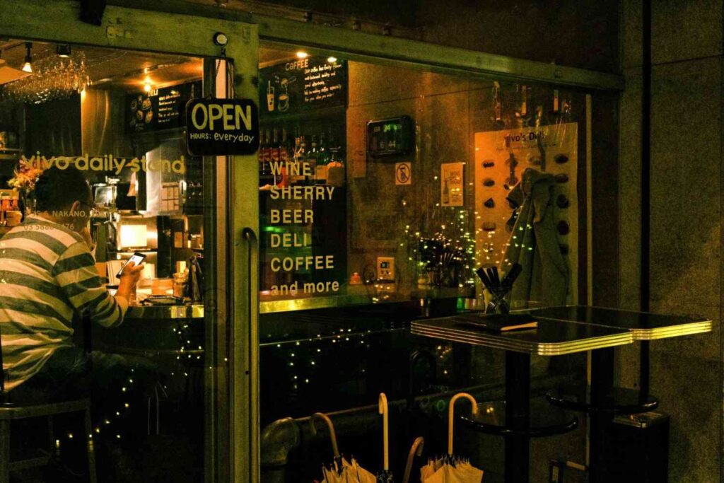 Illuminated by warm amber lighting, the glass storefront of "vivo daily stand" invites passersby with a window display listing offerings such as wine, sherry, beer, and coffee. Inside the cozy interior, a lone patron wearing a striped shirt sits at the counter, engrossed in his smartphone.