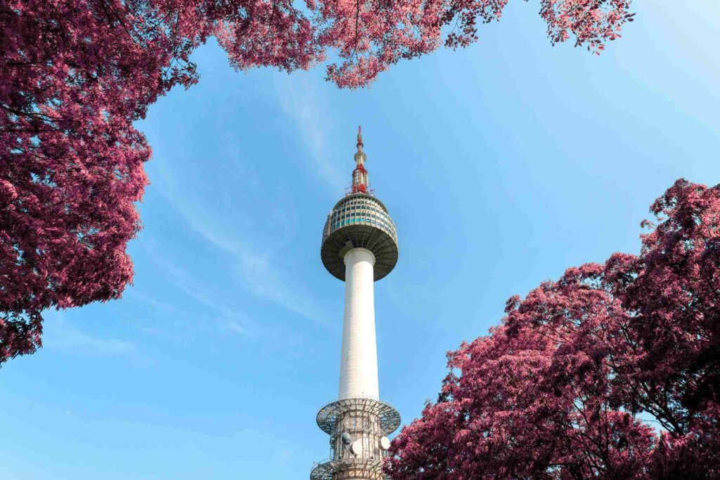 This low-angle, wide-shot image showcases the N Seoul Tower, a distinctive white structure with a rounded observation deck, soaring into a clear blue sky.