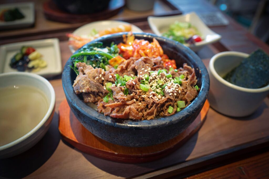 This close-up, high-angle image features a Korean dish of sliced marinated beef, likely Bulgogi served over rice in a black stone bowl, topped with sesame seeds and chopped scallions.