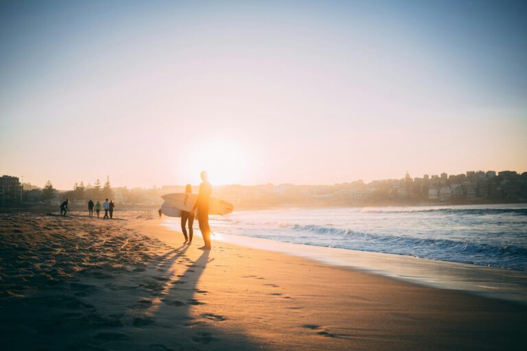 Bathed in the warm glow of a low sun, two surfers carrying boards stand on the sandy shore, casting long shadows across the beach. Gentle waves wash onto the coast to the right, while a hazy city skyline frames the background of this peaceful seaside scene.