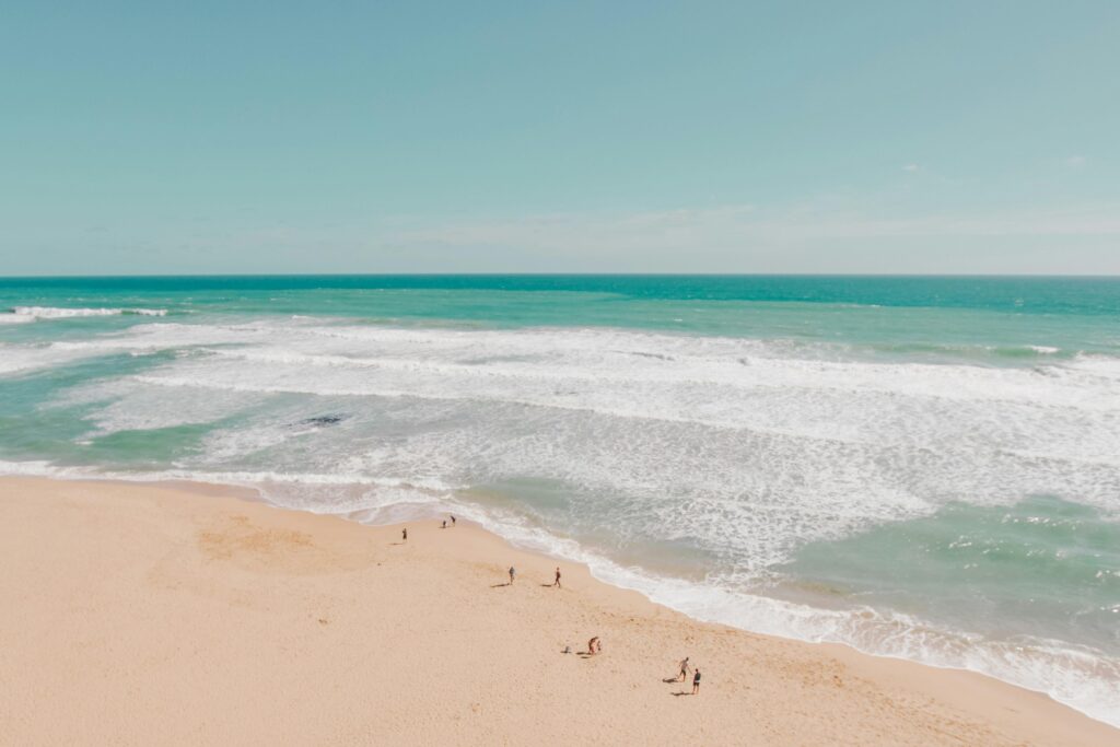 This high-angle shot captures a vast expanse of turquoise ocean rolling onto a pristine beige beach, with layers of white foamy waves stretching across the frame. Tiny figures of people scattered along the shoreline highlight the immense scale of the coastal landscape under a bright, clear sky.