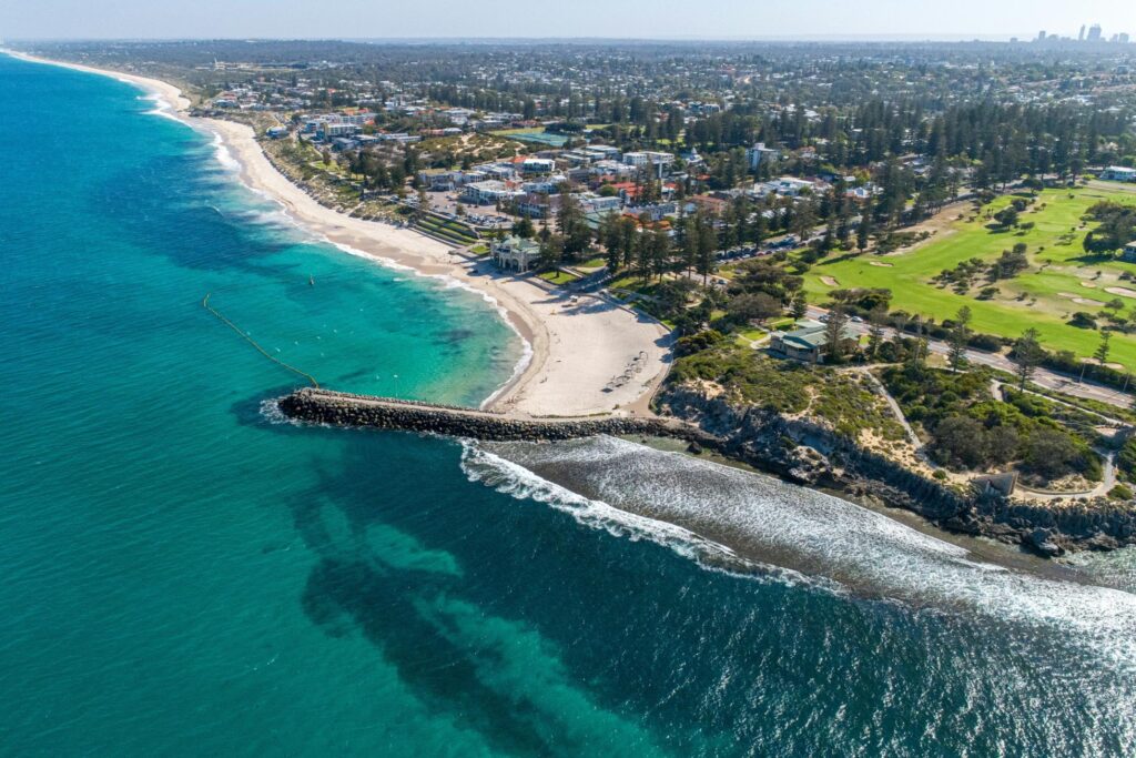 This aerial photograph captures a sweeping coastline where vibrant turquoise waters meet a sandy beach, featuring a prominent stone breakwater extending into the ocean. Inland, lush green parklands and residential areas border the shore, leading the eye toward a distant city skyline on the horizon.