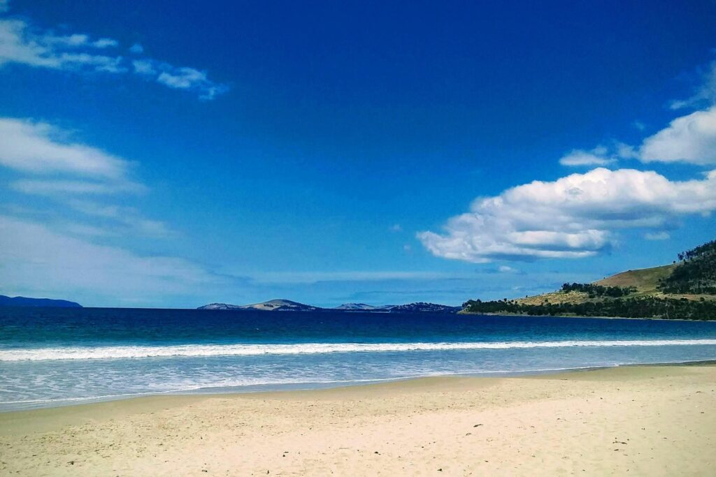 Under a vibrant blue sky dotted with fluffy white clouds, gentle waves roll onto a wide stretch of sandy beach. To the right, green rolling hills slope down towards the water, framing a view of distant landmasses on the horizon.