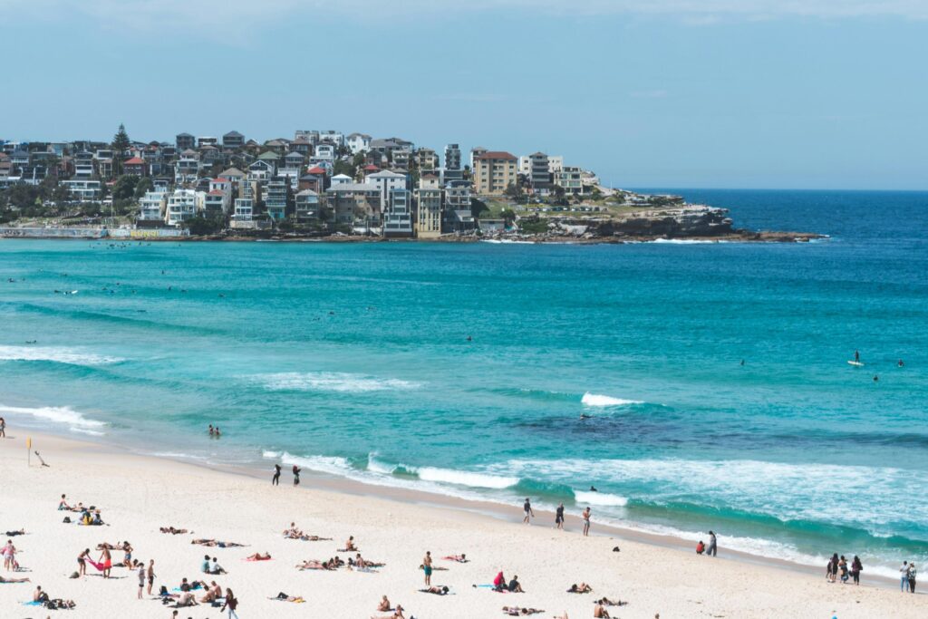 Crowds of people relax on the white sands and wade in the vibrant turquoise waters of a bustling beach. A dense cluster of multi-story buildings sits atop a rocky headland in the background, overlooking the ocean under a clear blue sky.