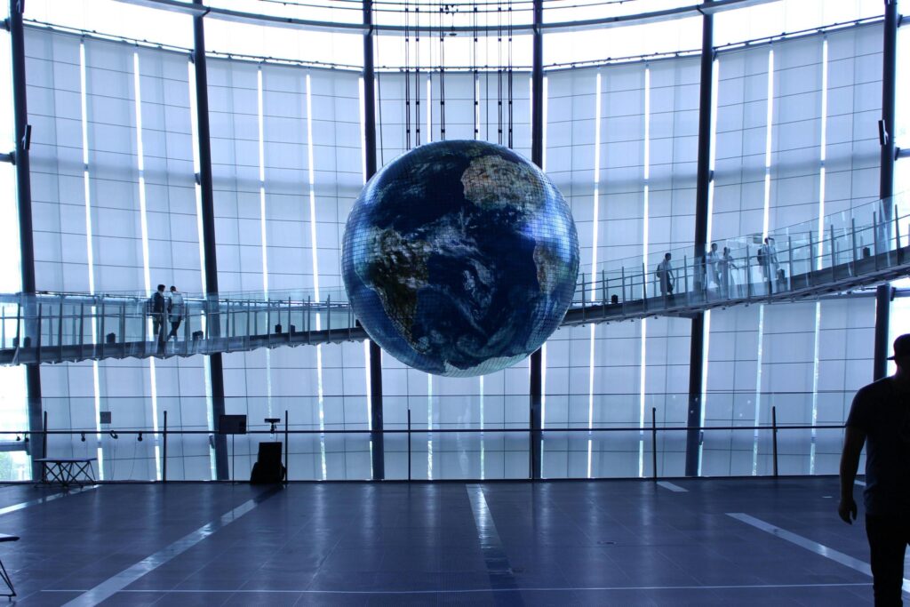 A massive, high-resolution globe hangs suspended in the center of a large, modern hall with translucent glass walls. Pedestrians walk along a curved glass walkway that encircles the floating sphere, highlighting the scale and futuristic design of the space.