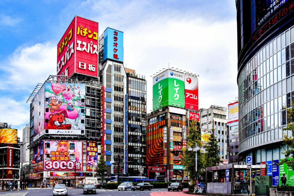 This vibrant street scene captures a bustling Japanese cityscape filled with multi-story buildings covered in colorful, high-contrast advertisements and neon signs. Cars navigate a wide intersection in the foreground, surrounded by a dense urban environment that includes visible landmarks like a McDonald's and various entertainment centers.