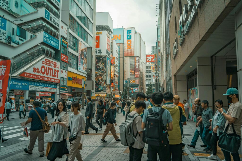 This image depicts a crowded pedestrian street in Akihabara, Japan, flanked by multi-story buildings adorned with vibrant electronics advertisements and anime-style billboards. Numerous people with backpacks and casual wear navigate the walkway, while signs for "Sofmap," "Laox," and "Akihabara Station" are visible in the background.