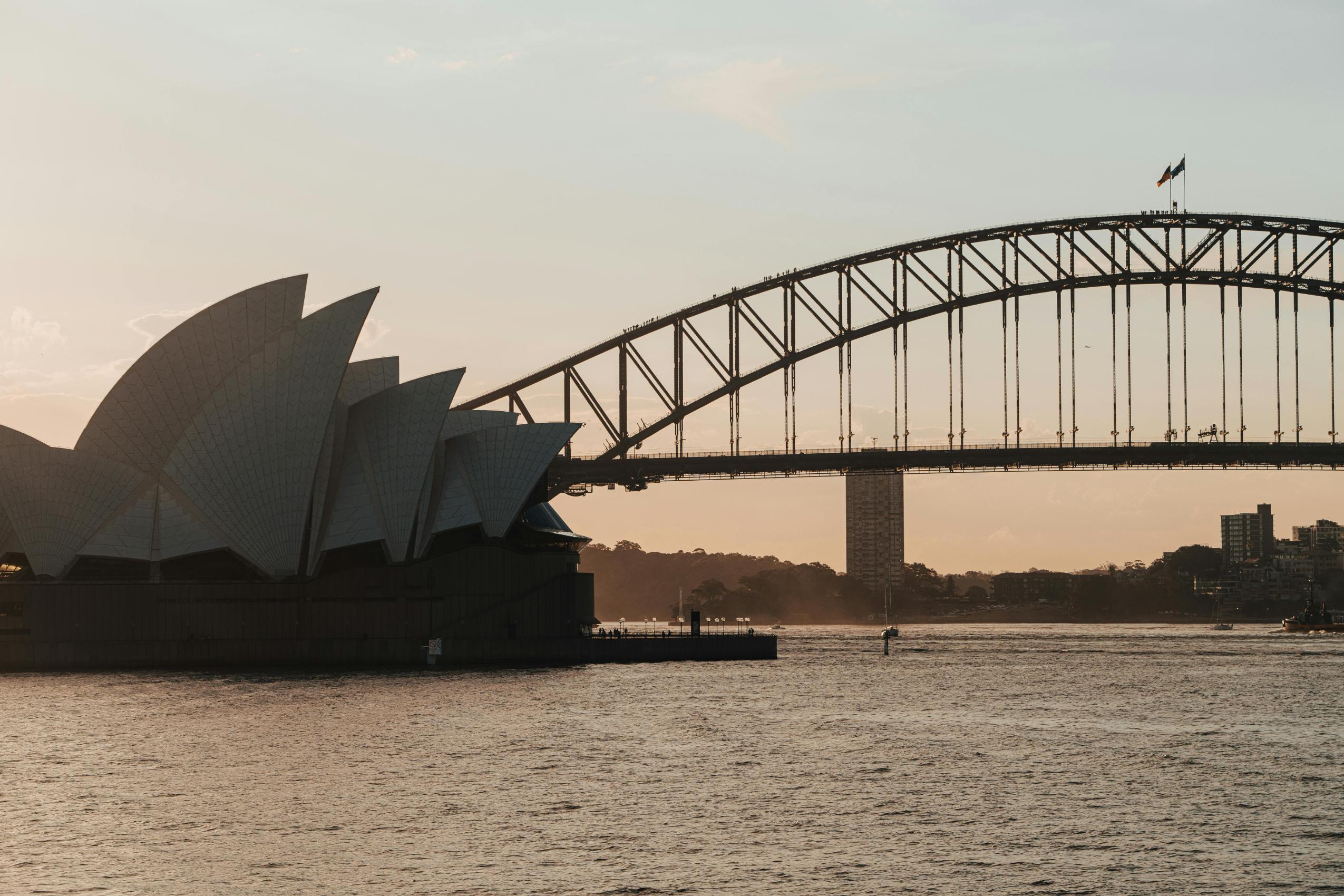 The image captures a scenic view of the iconic Sydney Opera House with its distinctive white sails, situated alongside the sweeping steel arch of the Sydney Harbour Bridge. Bathed in the warm, golden light of sunset, these famous landmarks form a striking silhouette against the shimmering waters of the harbor.