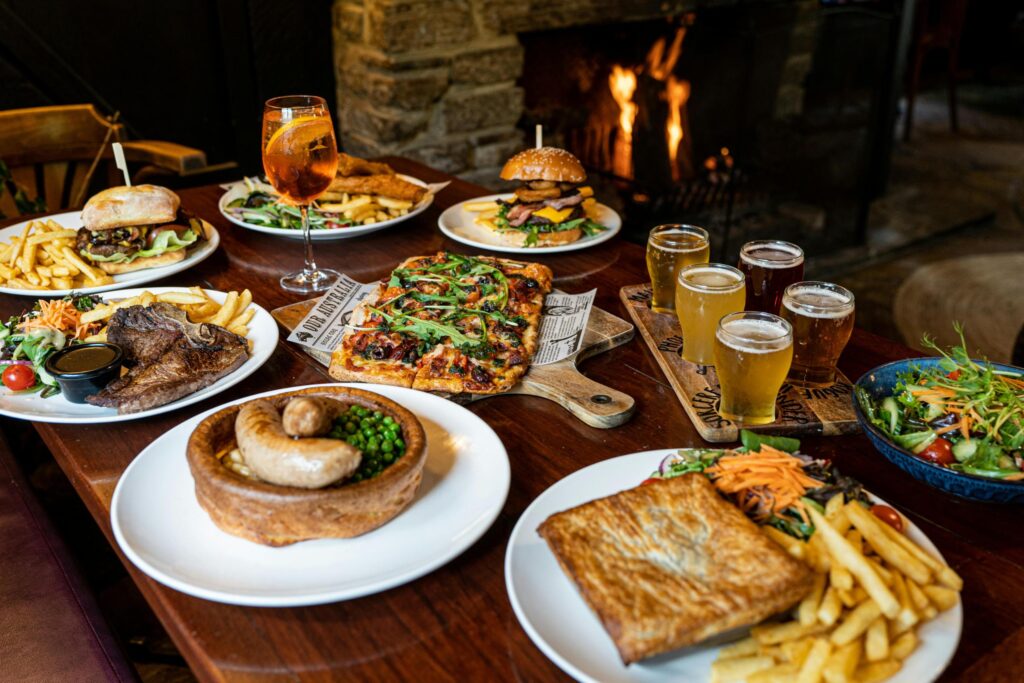 A rustic wooden table is laden with a bountiful feast of pub classics, including burgers, pizza, steak, savory pies, and sausages, all served with generous portions of fries and salad. The cozy atmosphere is enhanced by a flight of beers and an orange cocktail, while a roaring fire in the stone hearth background adds warmth to the inviting scene.