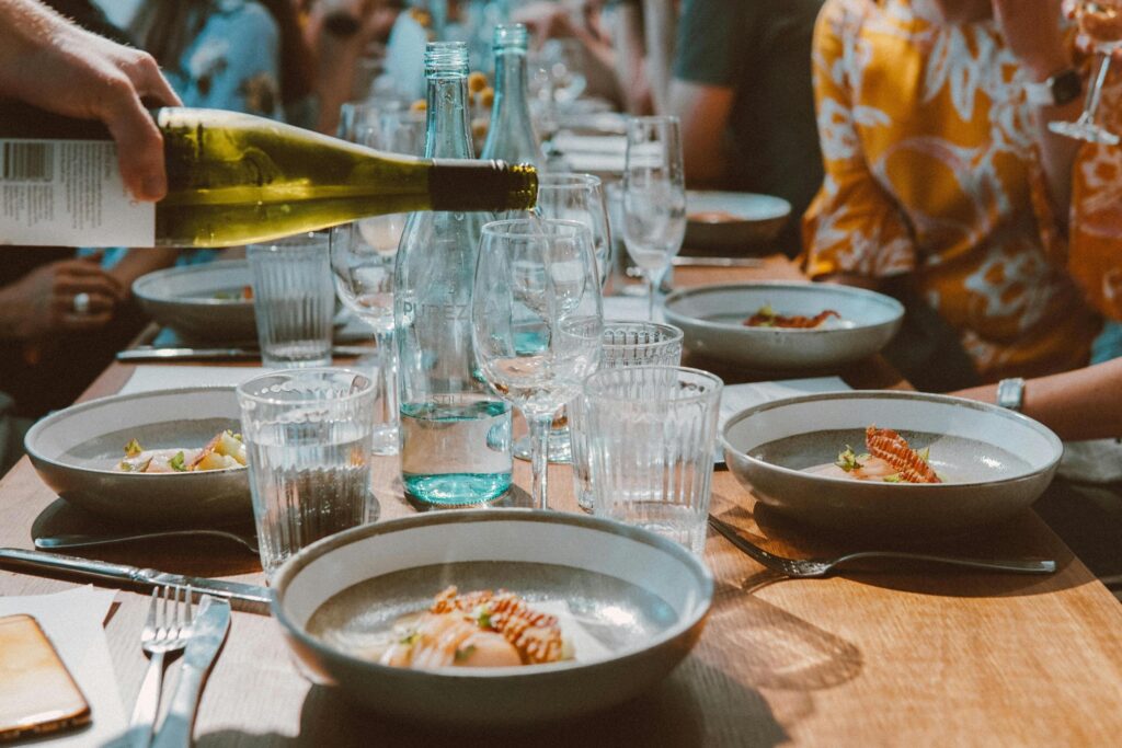 A hand pours white wine into a glass at a sun-lit dining table set with modern ceramic bowls containing elegantly plated appetizers. The scene captures a lively social gathering with sparkling glassware, a bottle of still water, and guests visible in the background enjoying their meal.
