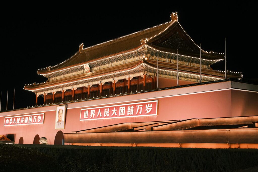 This illuminated nighttime photograph depicts the grand Tiananmen Gate, featuring towering red walls and a traditional double-eaved roof outlined in glowing lights. A large portrait sits centrally above the main archway, flanked by banners with white Chinese characters, standing out starkly against the dark night sky.