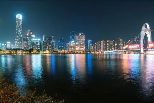 This vibrant nighttime cityscape features a skyline of towering, illuminated skyscrapers alongside a distinctive suspension bridge with a curved arch. The dazzling lights from the modern architecture reflect vividly across the calm surface of the wide river, creating shimmering streaks of color in the water.