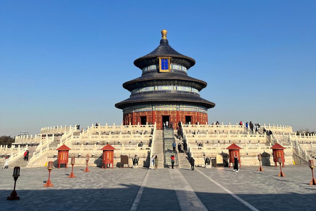 The iconic Hall of Prayer for Good Harvests stands centrally in the frame, displaying its three-tiered blue roof and ornate red woodwork atop a grand, multi-level white marble terrace. Below the temple, a paved plaza featuring red ceremonial stands bustles with visitors ascending the stairs, highlighting the scale of this historic site against a clear blue sky.