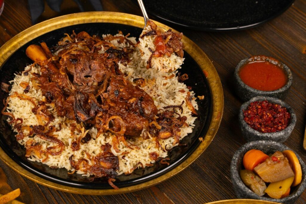 A generous serving of spiced rice and tender roasted meat sits on a gold-rimmed black plate, garnished heavily with crispy fried onions. Next to the main dish, a spoon lifts a portion of the food, while small stone bowls containing red sauces and pickled vegetables provide colorful accompaniments.