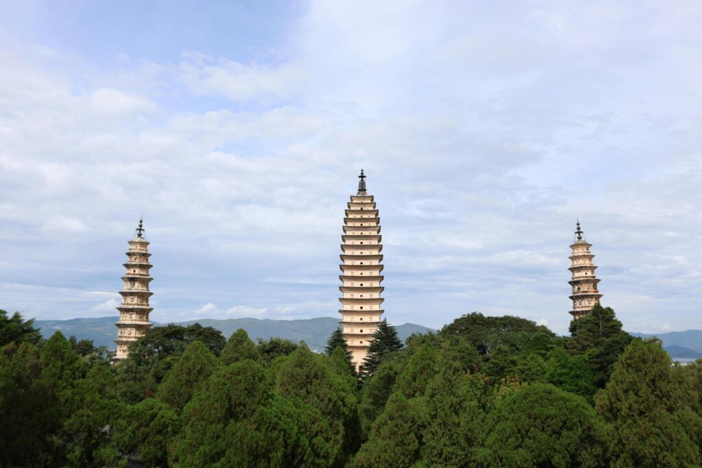 Three tall, multi-tiered pagodas rise majestically above a dense canopy of green trees, with the central tower standing significantly higher than the two flanking structures. The scene is set against a backdrop of distant, soft-hued mountains and a cloudy sky, emphasizing the height and serenity of the ancient architecture.