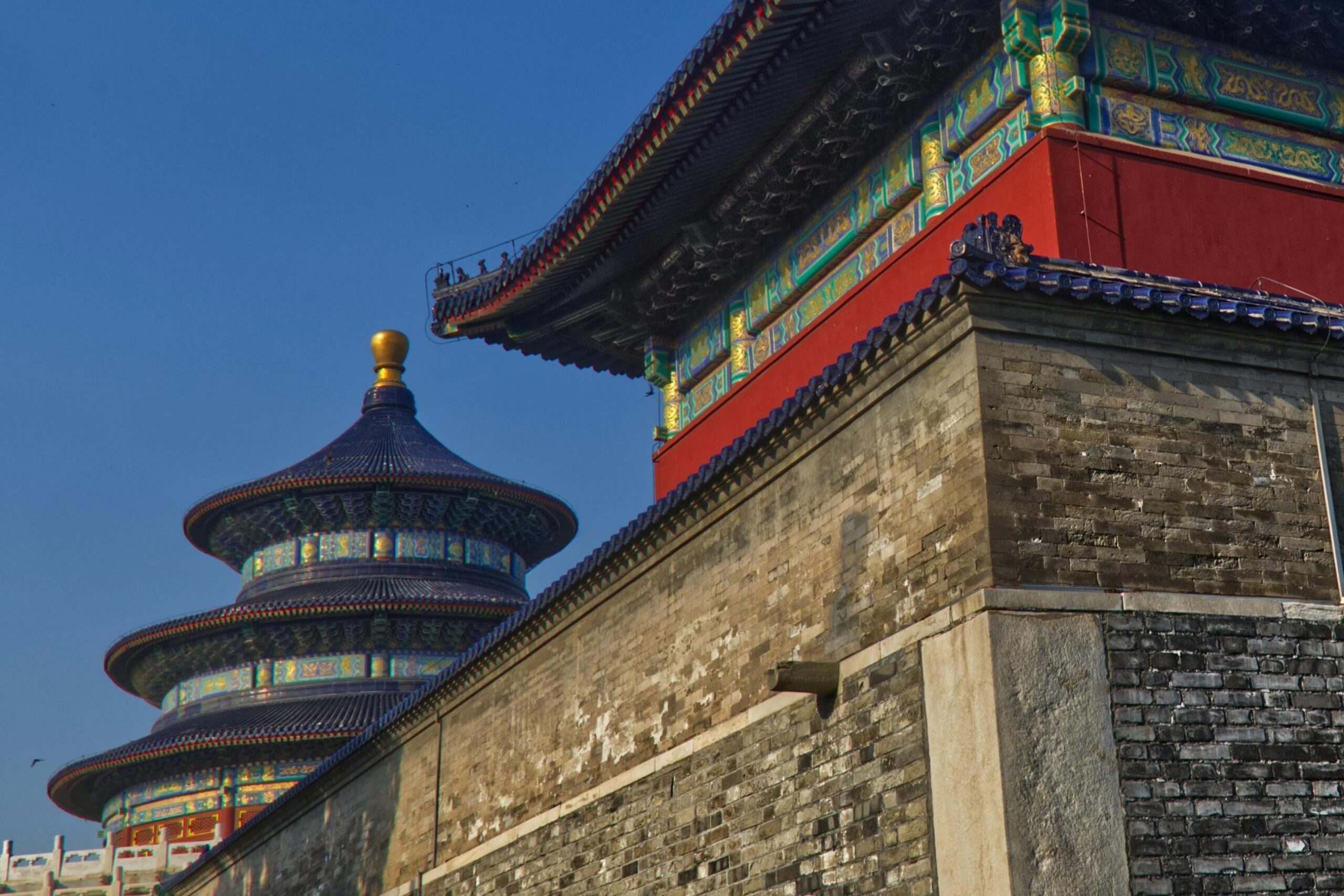 This image captures a striking low-angle view of the Temple of Heaven, featuring a weathered grey brick wall in the foreground that rises to meet an ornate roofline painted in vibrant red, green, and blue. Behind this structure, the iconic triple-tiered Hall of Prayer for Good Harvests stands out against a clear sky, distinguished by its circular shape, deep blue tiles, and golden finial.