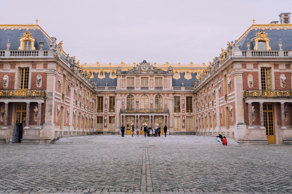 This image depicts the opulent Marble Court of the Palace of Versailles, characterized by its symmetrical red brick and cream stone facade adorned with lavish gold accents and statues. A vast cobblestone courtyard stretches out in the foreground where small groups of visitors gather, emphasizing the grand scale of this historic royal residence.