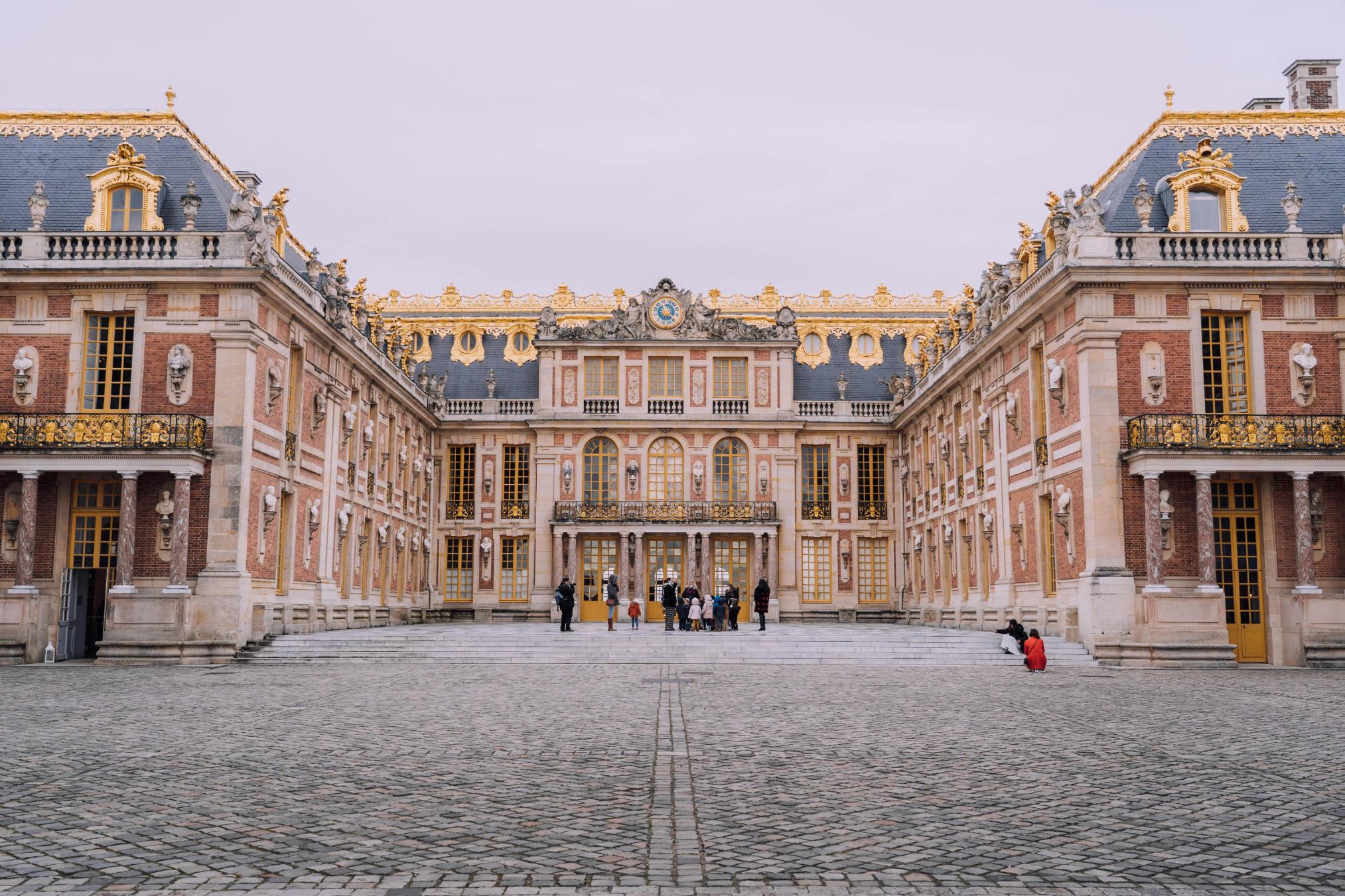 This image depicts the opulent Marble Court of the Palace of Versailles, characterized by its symmetrical red brick and cream stone facade adorned with lavish gold accents and statues. A vast cobblestone courtyard stretches out in the foreground where small groups of visitors gather, emphasizing the grand scale of this historic royal residence.