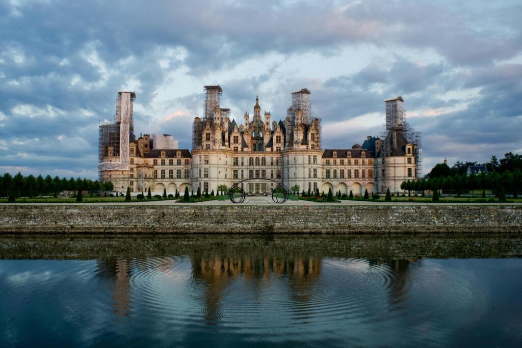 This image captures the majestic Château de Chambord, characterized by its elaborate roofline of turrets and chimneys that is mirrored in the calm waters of the foreground moat. The scene is set against a dramatic, cloudy sky that enhances the grandeur of the Renaissance architecture, which currently shows signs of restoration on its outer towers.