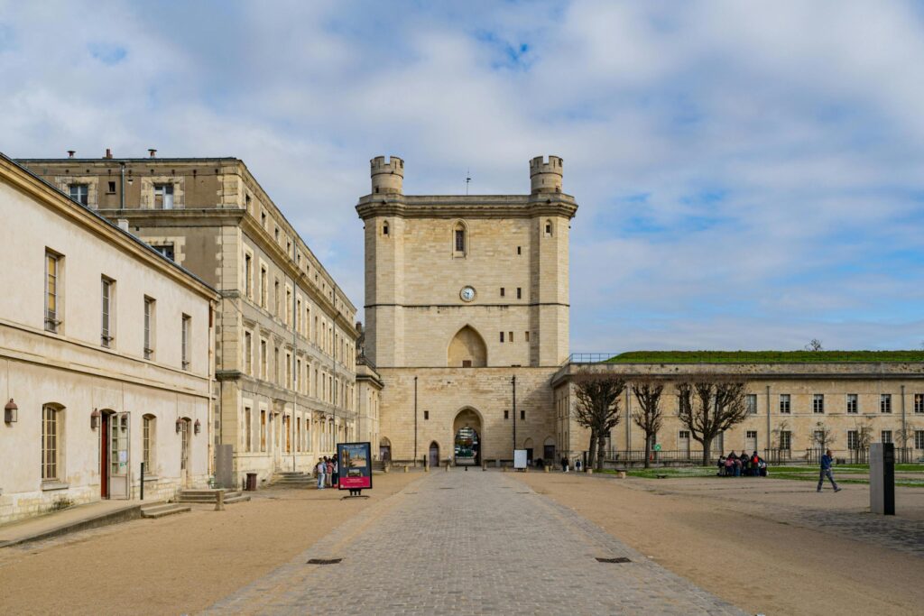 This image depicts the imposing entrance of a historic stone fortress, dominated by a tall, square tower featuring a clock face and corner turrets. A wide, paved pathway leads through the courtyard, flanked by long, multi-story buildings where visitors stroll under a bright, cloudy sky.
