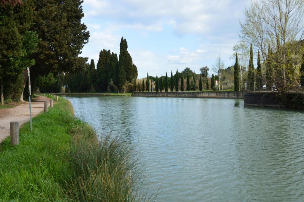 This serene landscape features a calm, wide canal bordered on the left by a dirt path and lush green vegetation. On the opposite bank, a distinctive row of tall, slender cypress trees stands along a concrete wall under a bright, partly cloudy sky.