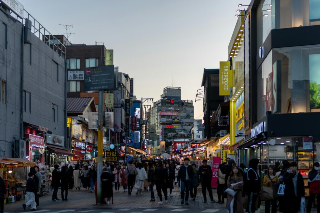 Crowds of pedestrians stroll through a bustling shopping district lined with densely packed multi-story buildings and street-level vendors. The scene is vibrant with illuminated storefronts and vertical signage in Korean and English, highlighting brands like "SKINFOOD" against the soft light of dusk.