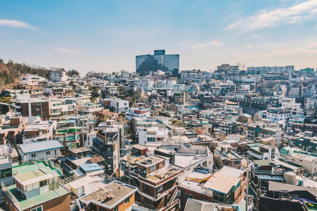 Crowds of pedestrians stroll through a bustling shopping district lined with densely packed multi-story buildings and street-level vendors. The scene is vibrant with illuminated storefronts and vertical signage in Korean and English, highlighting brands like "SKINFOOD" against the soft light of dusk.
