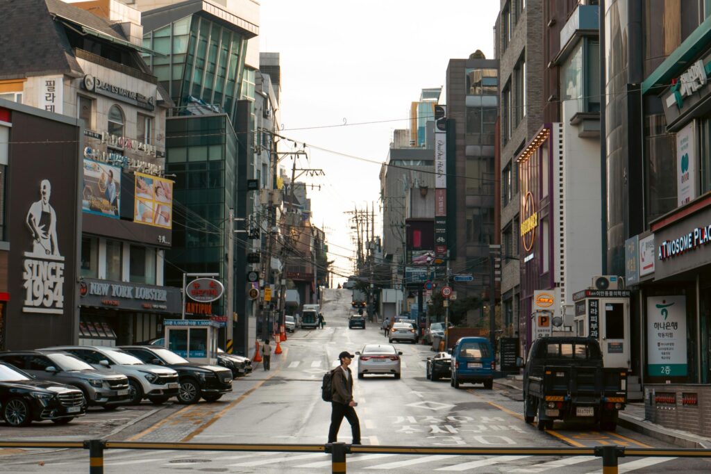 A narrow, paved road descends through a quiet neighborhood of traditional Korean houses featuring ornate brick walls and curved, tiled roofs. In the background, this historic architecture contrasts with a modern city skyline, highlighted by the distinct silhouette of the N Seoul Tower atop a distant hill.