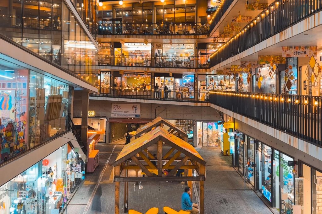 This vibrant image captures a multi-level, open-air shopping complex at night, glowing with warm string lights along its spiraling walkways. Bright glass storefronts fill the tiered balconies, overlooking a central courtyard where visitors stroll near a wooden pavilion.