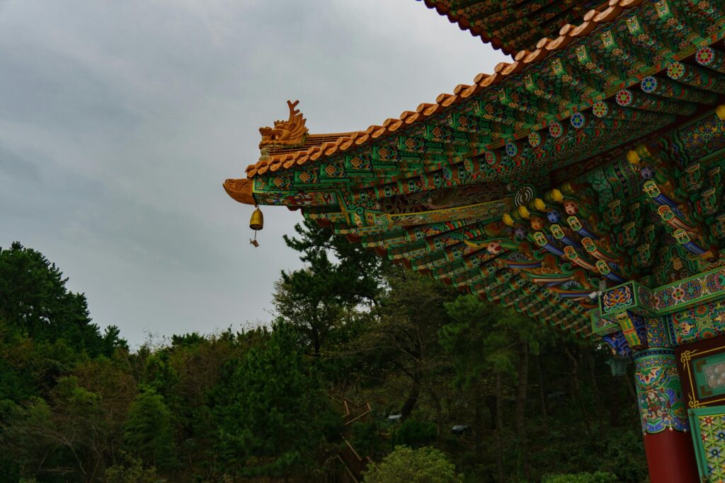 The image captures the intricate underside of a traditional Korean roof, featuring elaborate wooden brackets painted with vibrant green and red patterns known as Dancheong. A dragon head ornament sits atop the corner, from which a small wind chime hangs against a backdrop of dense, dark green forest and a gray sky.